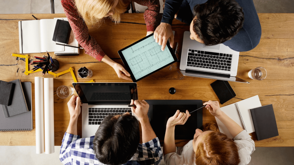 Ein tisch von oben auf dem mehrere Laptops und Tablets stehen und an dem ein Team aus Männern und Frauen zusammenarbeitet.