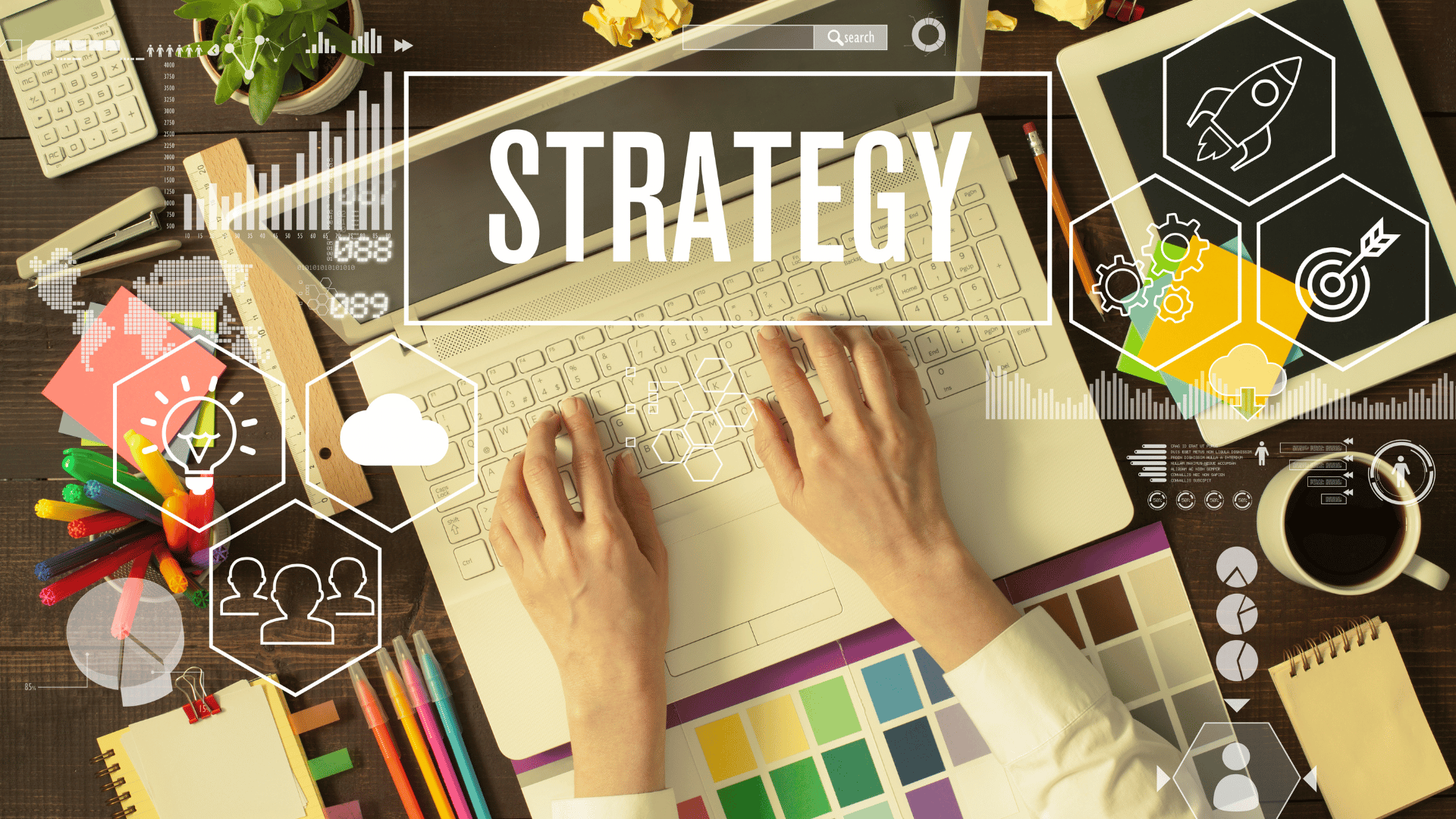 Der Blick geht von oben auf einen Schreibtisch. Zwei Hände schreiben auf einem Laptop, um das Gerät herum liegen Stifte und Notizen. Über dem Laptop schwebt das Wort "Strategy"