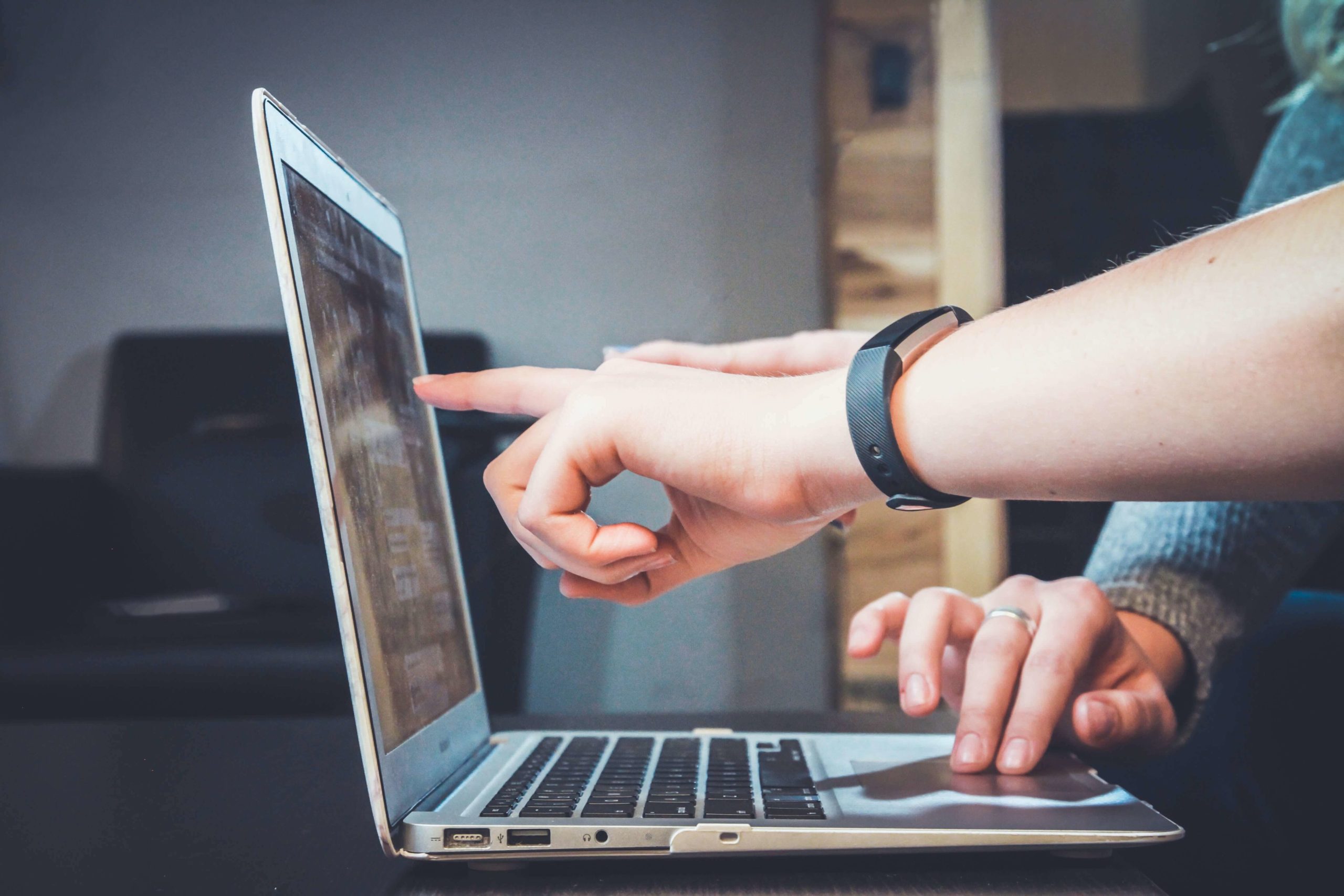 Auf dem Bild ist ein Laptop zu sehen, sowie eine Hand die etwas auf dem Laptop zeigt und eine andere Hand die auf dem Laptop tippt.
